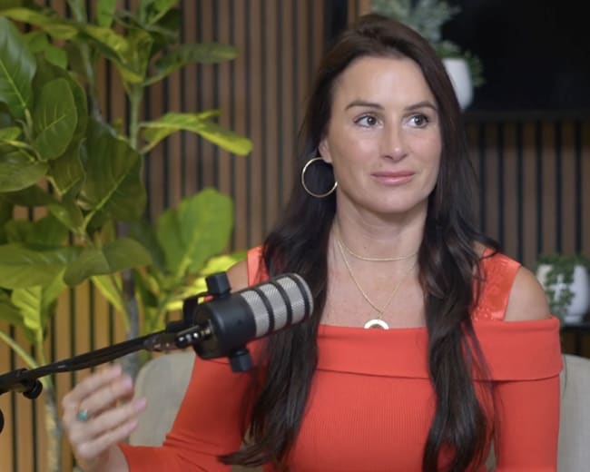 A woman with long dark hair is sitting in front of a microphone, wearing an off-the-shoulder red top, while gesturing with her hand amidst a backdrop of plants and wooden panels.