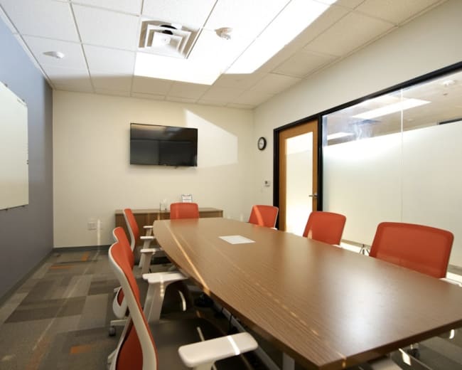 A modern meeting room features a long wooden table surrounded by white chairs, a flat-screen TV mounted on the wall, and a whiteboard.
