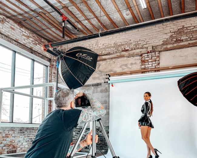 A photographer crouches behind a camera on a tripod, capturing a model posing in a shiny outfit against a light backdrop in a studio with exposed brick walls.