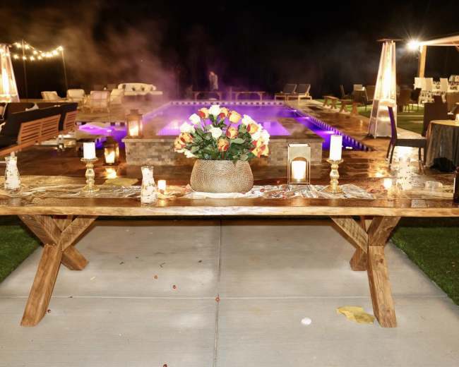 A large wooden table is set with a floral centerpiece and candles near a pool illuminated with purple lights at night.