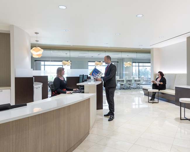 A man stands at a reception desk while a woman works on a computer, and another woman sits in a waiting area in a modern office lobby.
