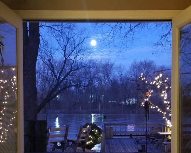 A view of a river with trees and a deck illuminated by lights, framed by open double doors under a moonlit sky.