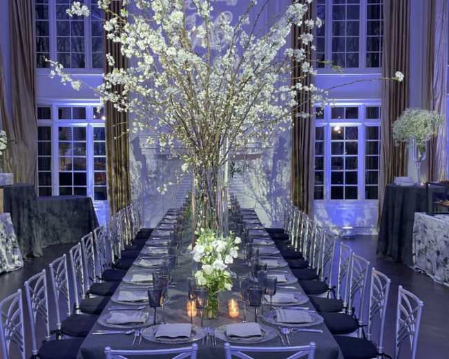 A long dining table is set in a spacious room with elegant decor, featuring tall floral arrangements and a warm glow from lighting.