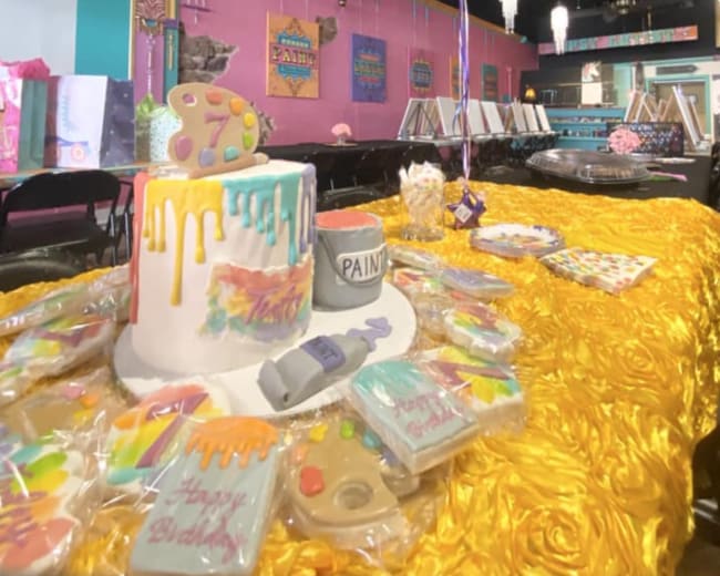 A vibrant party setup featuring a colorful cake, decorated cookies, and balloons, surrounded by party supplies in a bright venue.