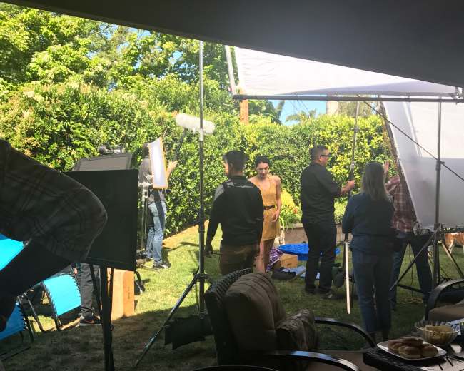 A film crew sets up equipment for a shoot in a backyard surrounded by greenery.