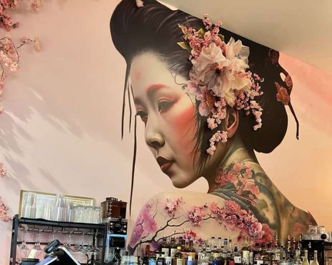 The image shows a bar with a large mural of a woman adorned with flowers and traditional Japanese elements, alongside a sake bottle and a glass on the counter.