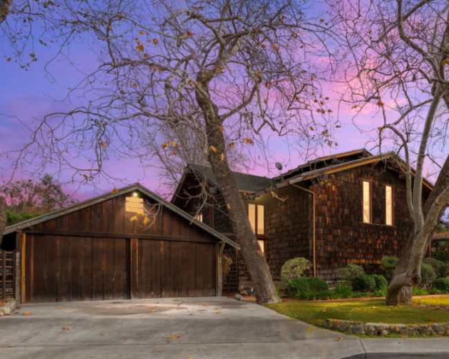 A two-story, wooden-shingled house with a garage is surrounded by bare trees and green lawn under a colorful twilight sky.