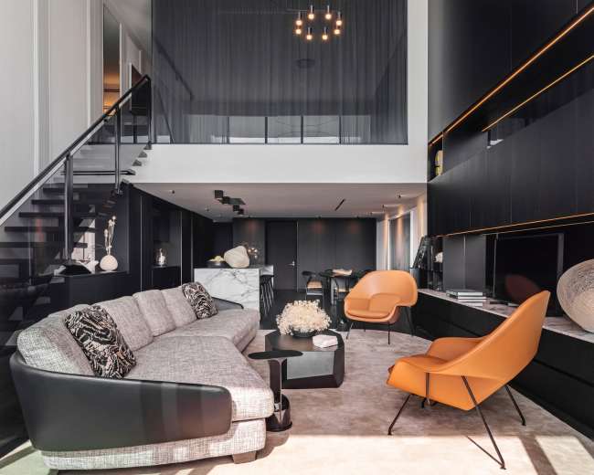 The image shows a modern living room with a gray sofa, an orange chair, a glass staircase, and a high ceiling featuring a geometric light fixture.