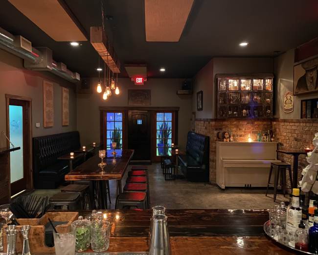 The interior of a dimly lit bar features a long wooden bar, black leather seating, and a brick wall with illuminated shelves.