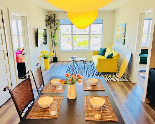 A modern apartment features a dining area with a table set for four, a living space with a yellow sofa, and large windows that provide natural light.
