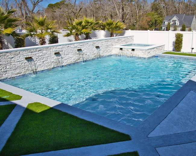 A rectangular swimming pool with a stone wall fountain, surrounded by clean landscaping and a spa area.