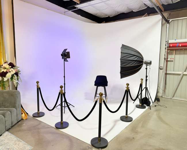 The image shows a photography studio setup with a white backdrop, a chair, lighting equipment, and a velvet rope barrier.