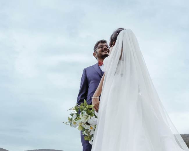 A bride in a flowing white dress holds a bouquet while standing with a groom in a suit under a cloudy sky.