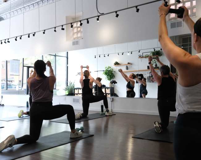 A group of women is engaged in a workout session in a fitness studio, using dumbbells and performing various exercises in front of a mirror.
