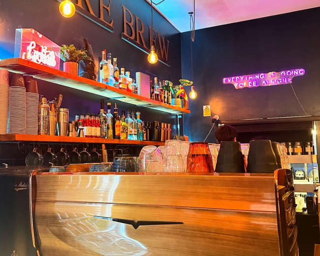 The image shows a coffee bar with shelves displaying various bottles and glassware, illuminated by warm hanging lights and a neon sign that reads "everything is going coffee alright."