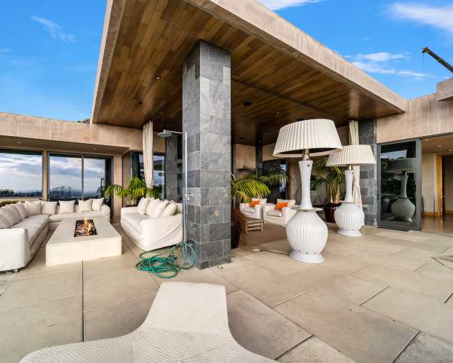 The image shows a modern outdoor patio area with large sofas, decorative floor lamps, and a view of the surrounding landscape.