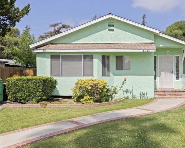 The image shows a light green single-story house with a front yard featuring a walkway and manicured bushes.