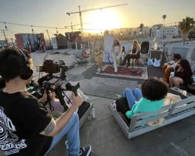 A film crew records a performance on a rooftop as the sun sets in the background, with several people seated in casual seating arrangements.