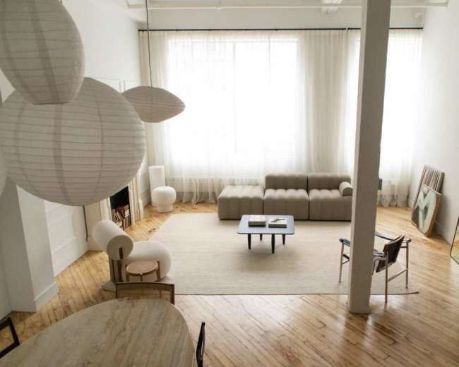 The image shows a modern, minimalist living space with a sectional sofa, a coffee table, and large windows allowing natural light to illuminate the room.