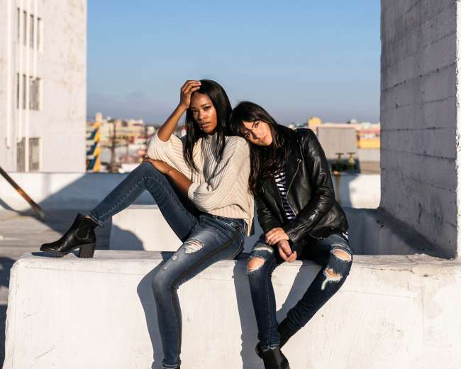 Two women sit on a concrete ledge wearing casual outfits, with one leaning back and the other resting her head on the first's shoulder, against a clear blue sky.