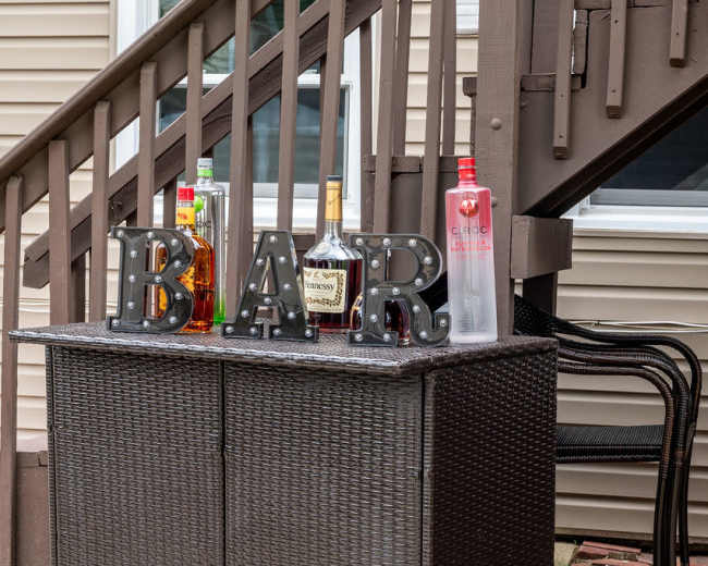 A wicker bar cart displays various liquor bottles alongside illuminated letters spelling "BAR."