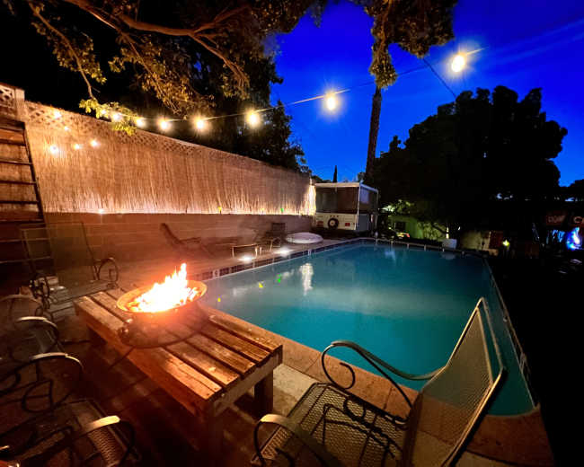 A swimming pool with a fire pit and surrounding seating area is illuminated by string lights in the evening.