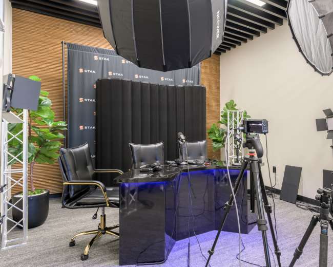 The image shows a well-equipped studio setup with a black desk, two chairs, and various cameras and lighting equipment arranged for a video recording.