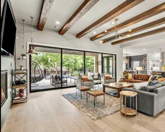 The image features a modern living room with wooden beams, a large sectional sofa, and a view of an outdoor patio through sliding glass doors.