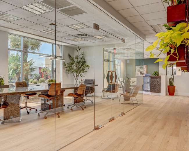 The image shows a modern office space with a glass partition, featuring a long conference table surrounded by chairs, and potted plants in the foreground.
