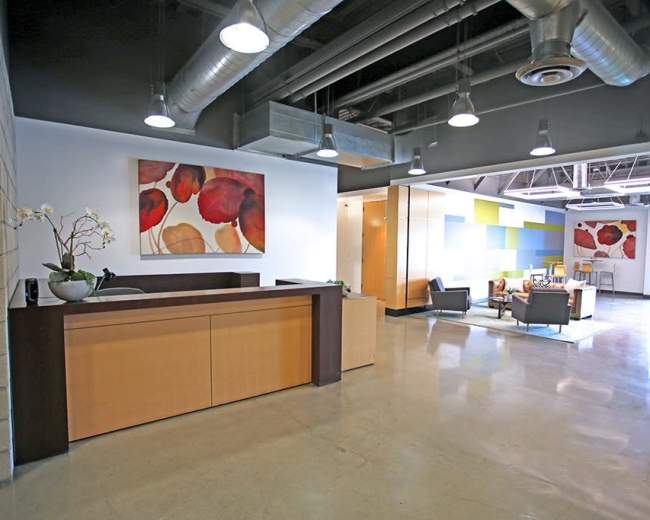 The image shows a modern office reception area with a wooden desk, colorful artwork on the walls, and a seating area in the background.