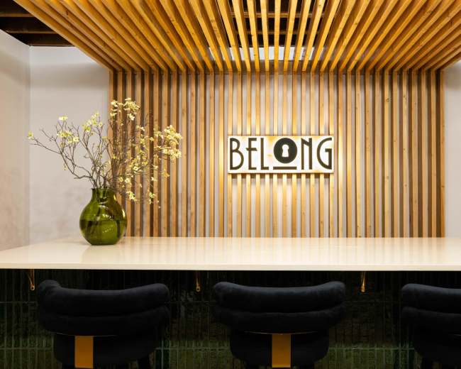 A modern interior with a wooden slatted wall featuring a sign that says "BELONG" above a sleek bar with dark cushioned stools and a green vase holding flowers.