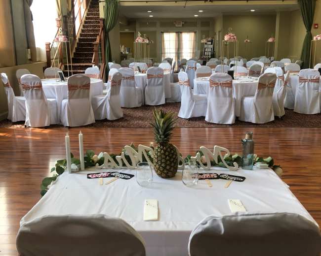 A banquet hall is set up for an event, featuring several tables covered with white tablecloths and decorated with rose-gold accents.