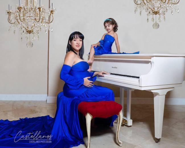 A woman in a blue gown sits at a white piano while a girl in a matching blue dress sits playfully on top of the piano.