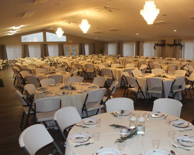A large banquet hall set up with numerous round tables, each covered with white tablecloths and arranged with plates and glassware.