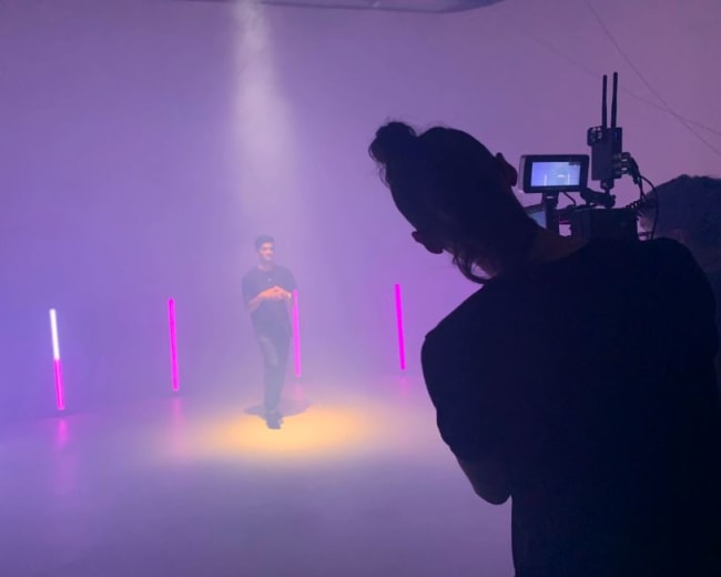 A person stands under a spotlight in a fog-filled room while being filmed by another person using a camera.