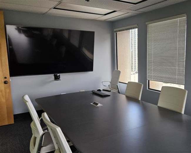 A conference room features a large black screen mounted on the wall, a rectangular table surrounded by white chairs, and windows with blinds.