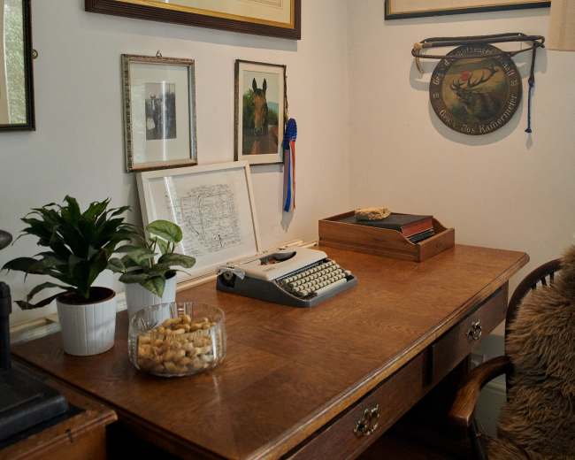 A wooden desk is positioned against a wall, surrounded by framed artwork and photographs, with a typewriter and decorative items on the surface.
