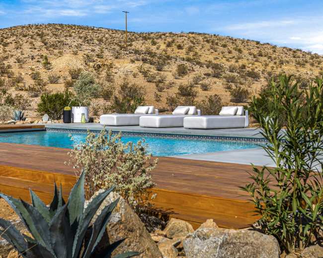 A modern swimming pool area surrounded by desert landscape, featuring lounge chairs and rocky terrain.