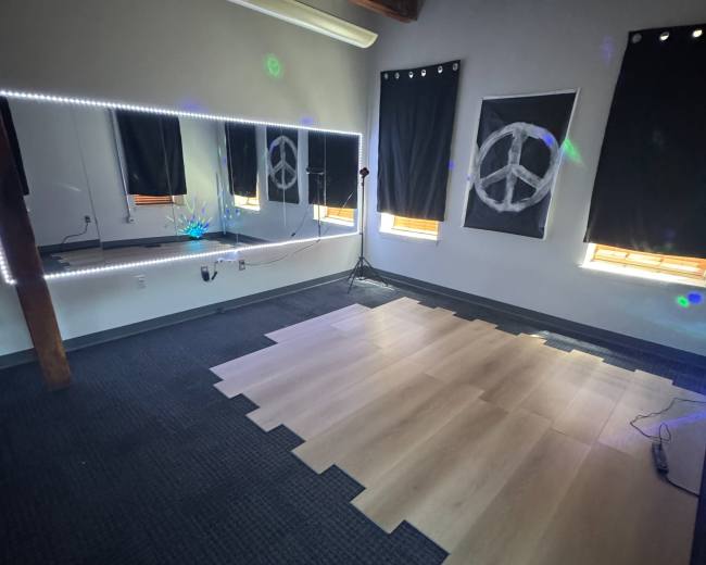 A spacious room with dark curtains, wooden beams, and partially laid light-colored flooring, featuring a mirrored wall with LED lights and peace symbol decorations.