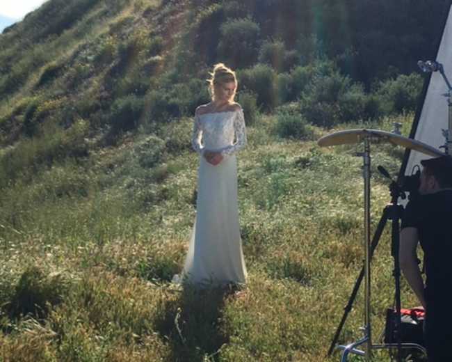 A woman in a long white dress stands on a grassy hillside while a photographer sets up equipment nearby.