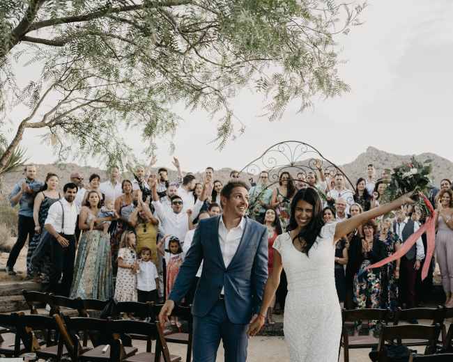 A newlywed couple walks hand in hand, smiling and celebrating, as their guests stand behind them, cheering in a desert landscape.