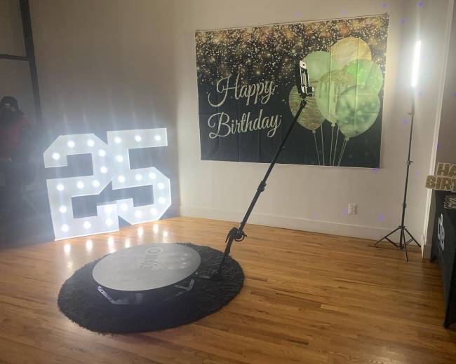The scene features a large illuminated "25" sign next to a backdrop of balloons and a banner that reads "Happy Birthday," with a photo booth setup in a well-lit room.