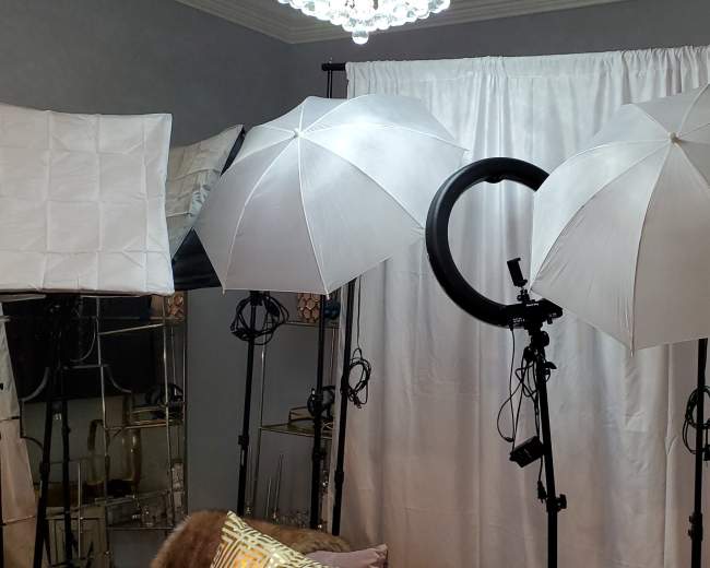 Three photography lighting setups, including softboxes and a ring light, positioned in front of a white backdrop in a room.