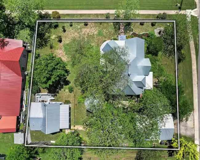 The image shows an aerial view of a residential property surrounded by green trees and grassy areas, with several nearby houses.