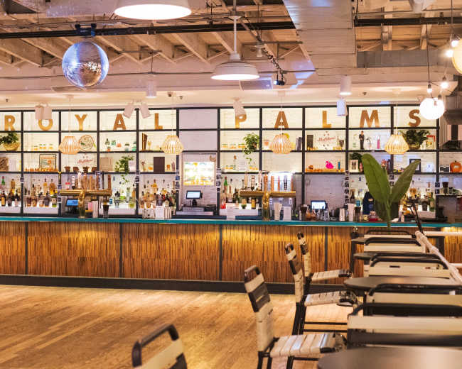 The image shows a well-organized bar area with a wooden counter, shelves displaying various bottles, and a large sign that reads "ROYAL PALMS."
