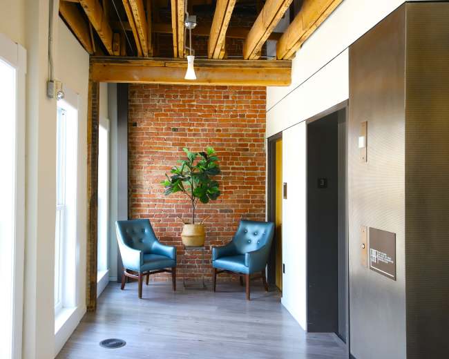 A hallway features two blue chairs beside a potted plant, with exposed brick walls and wooden beams above.