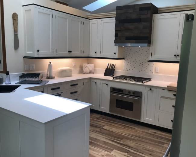 The image shows a modern kitchen with white cabinetry, a central island, stainless steel appliances, and wooden flooring.