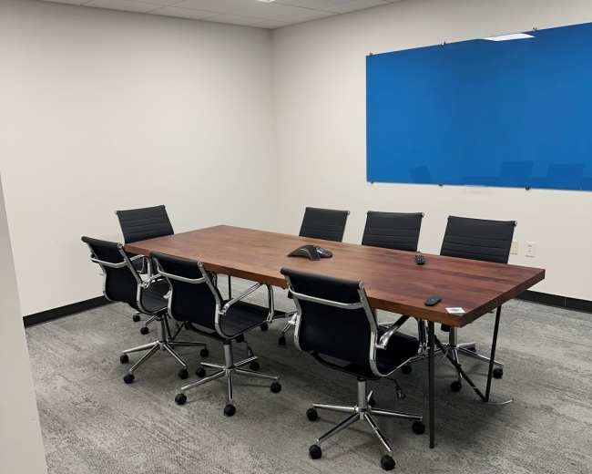 The image shows a conference room with a large wooden table surrounded by six black rolling chairs, and a blue wall in the background.