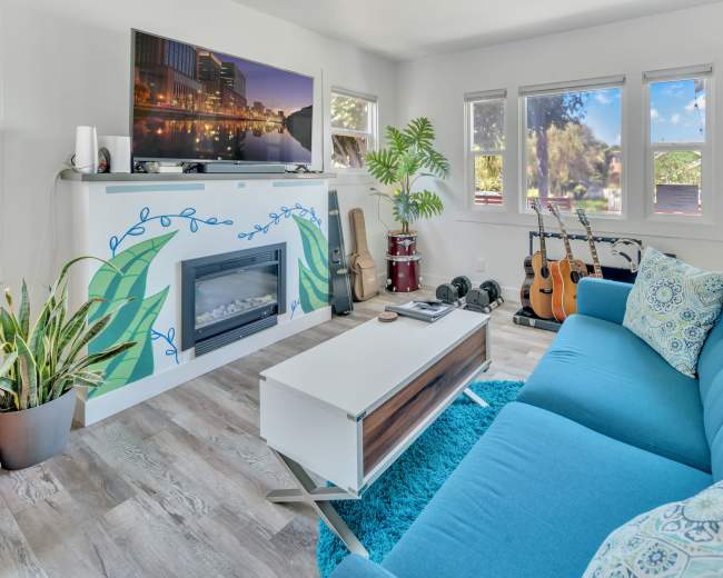 The image shows a bright living room featuring a blue couch, a wall-mounted television, a modern fireplace, and various musical instruments.