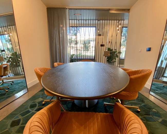 A round wooden table surrounded by four orange chairs is positioned in a room with large mirrors and glass doors leading to a garden.